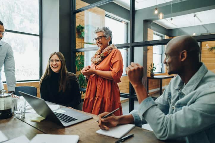 A smiling person stands up at a table with their enthusiastic coworkers.