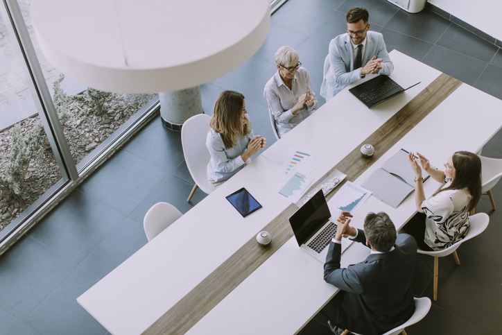 Business people sit on two sides of a long table in a modern office space with laptops and papers.